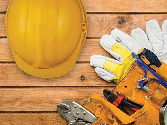 A bustling outdoor market scene with produce displayed in the foreground, including leafy greens and other vegetables. Two men stand behind the stall under blue tarpaulin, one smiling, suggesting a friendly and lively atmosphere.